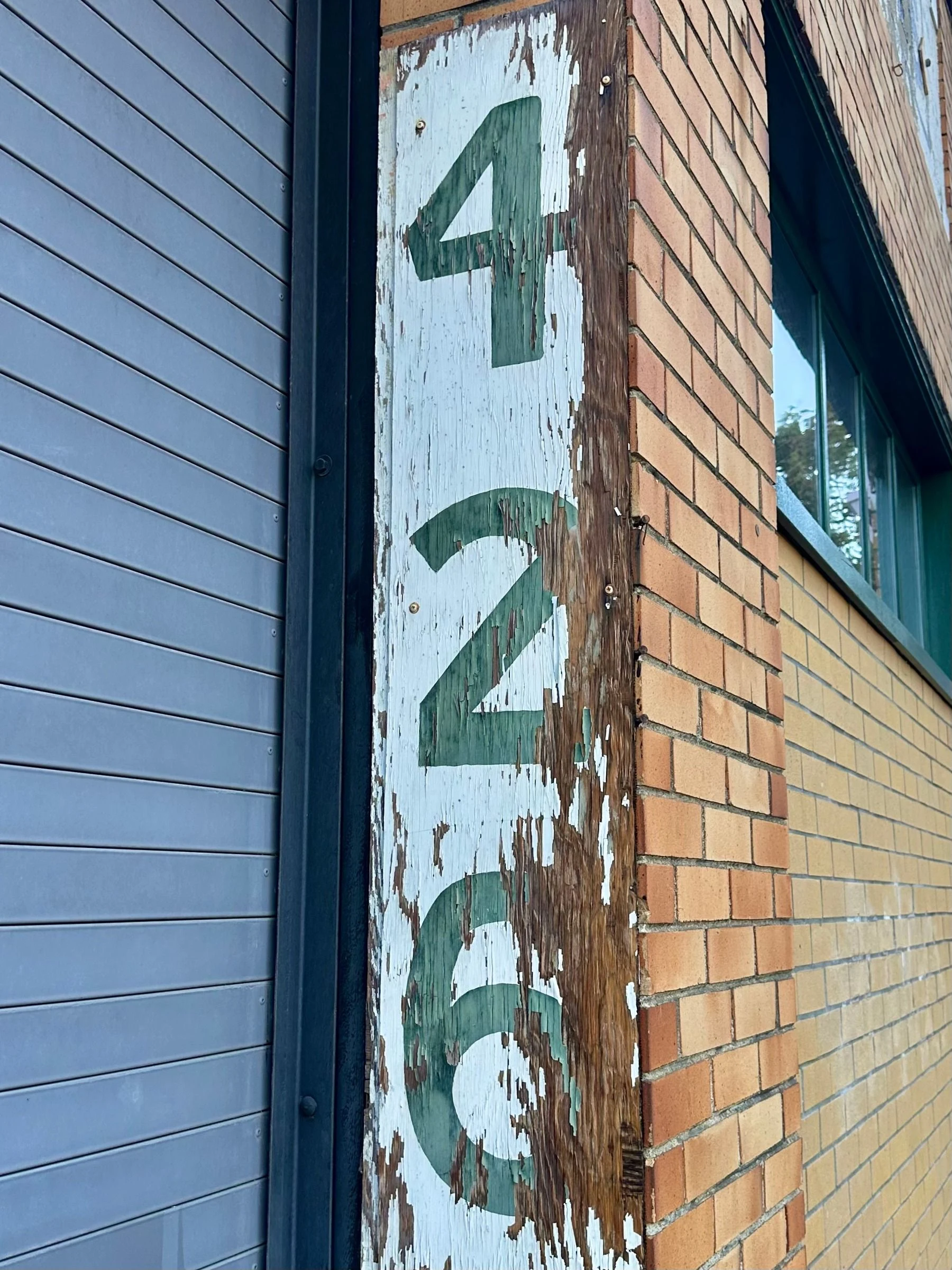 Green stenciled numbers 4, 2, 6 stacked vertically on a peeling white-painted wooden post between a gray roll-up door and a tan brick wall