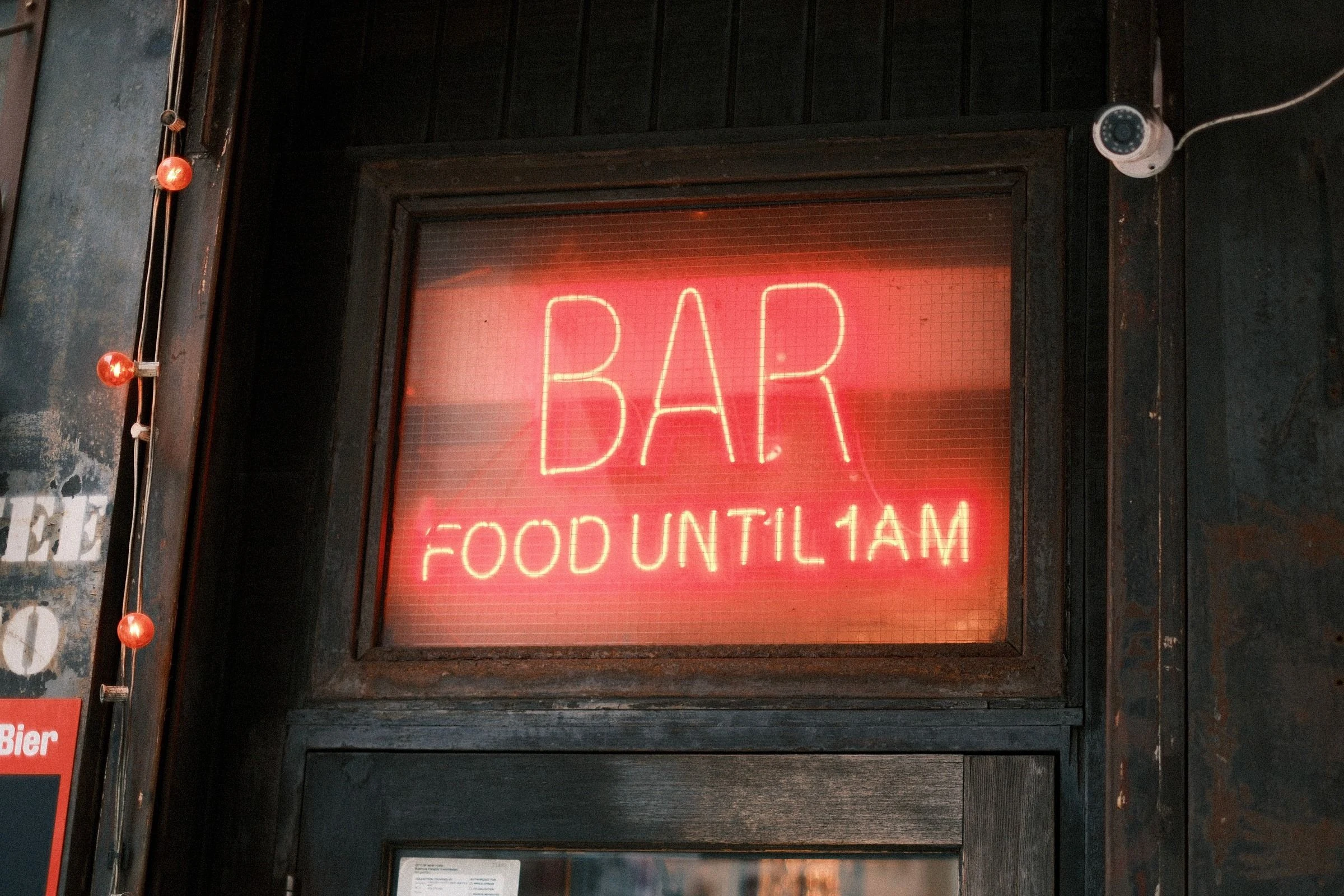 Red neon sign reading BAR and FOOD UNTIL 1AM glowing behind a wired-glass window above a weathered dark wooden door