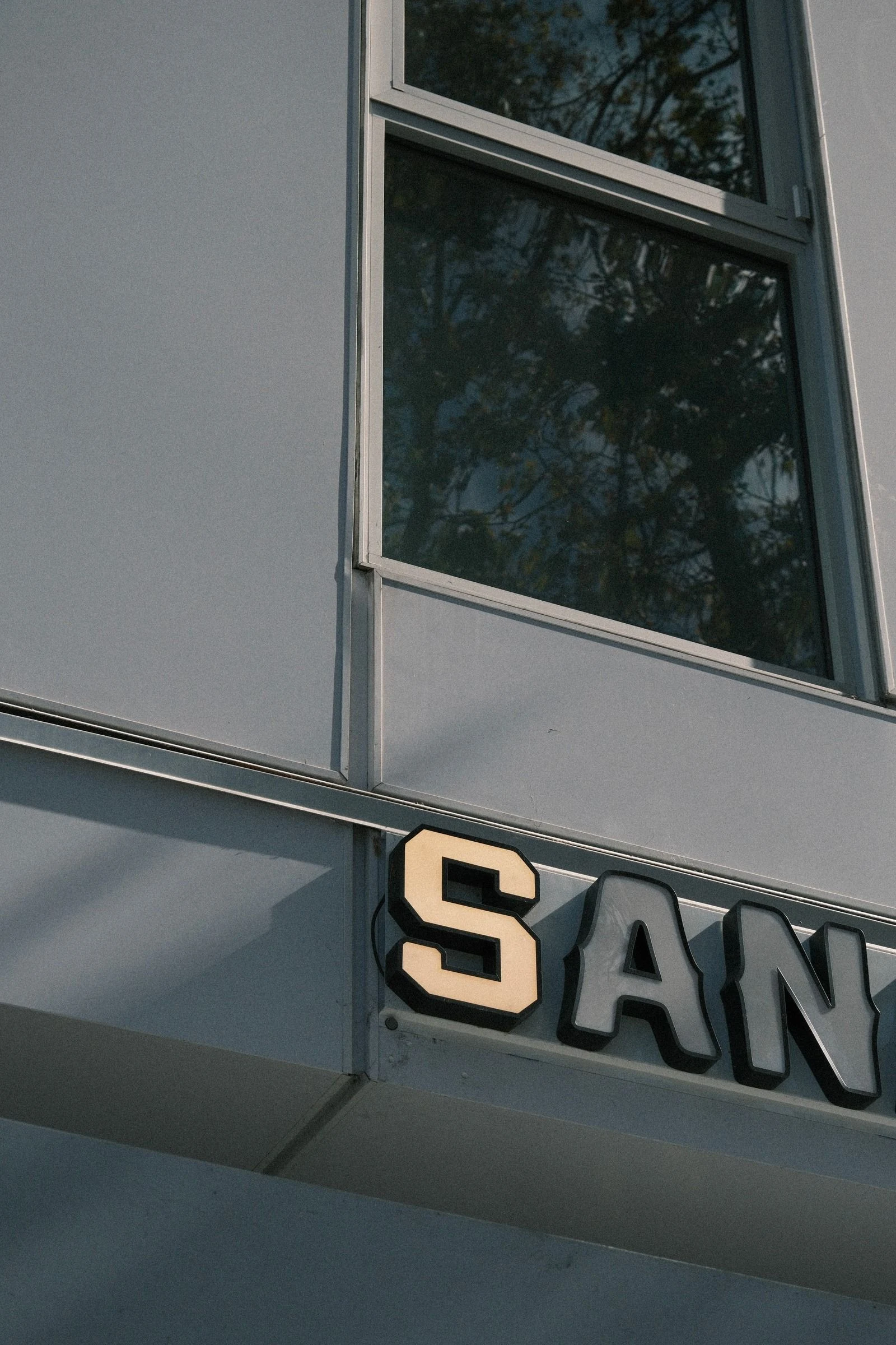 Dimensional block letters S, A, N mounted on a pale gray building facade below a window reflecting trees, with the S lit warm cream