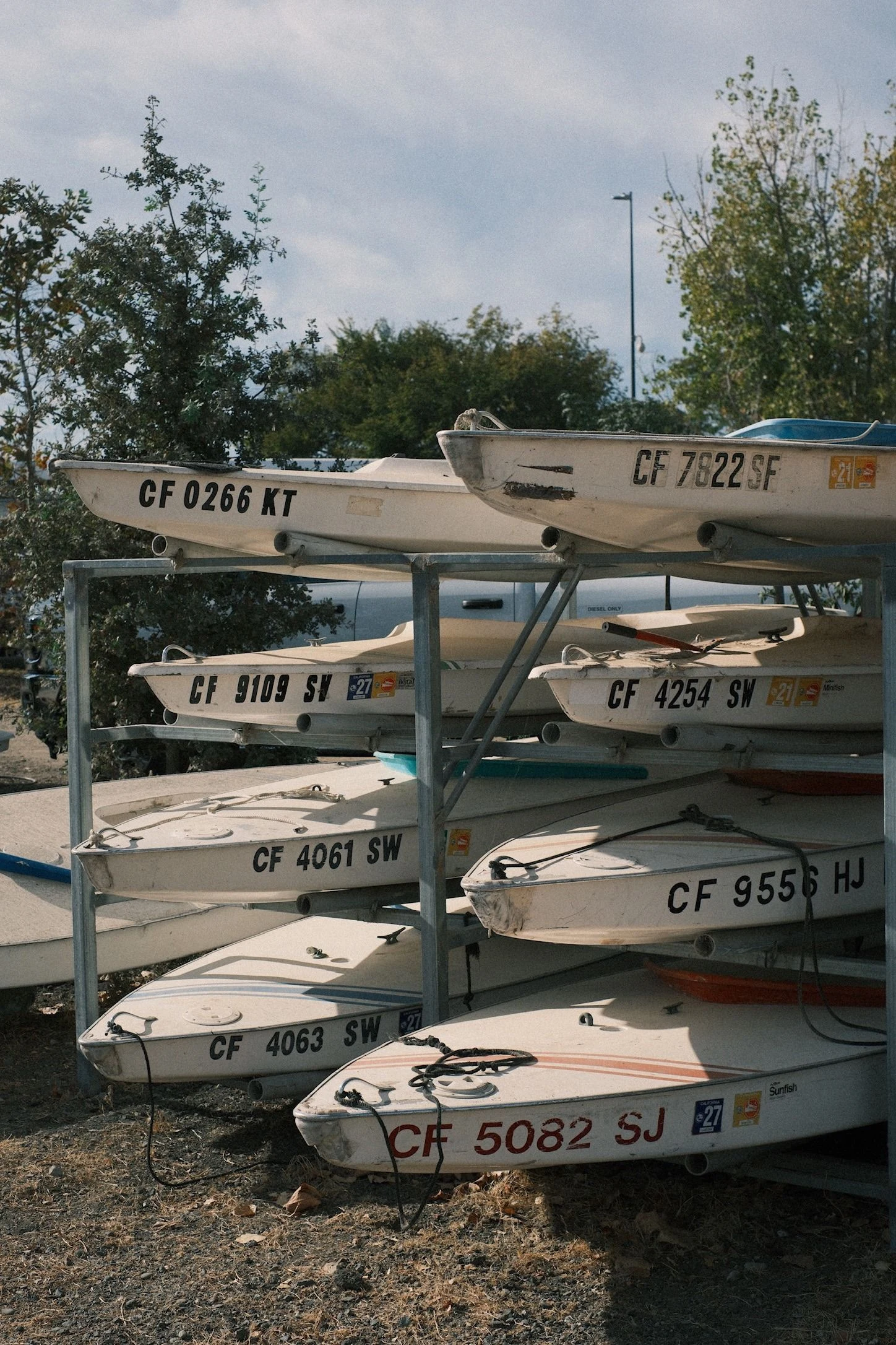 Rack of small white sailboat hulls stacked on a metal frame, each with a stenciled registration number like CF 0266 KT and CF 5082 SJ, under a cloudy sky with trees behind