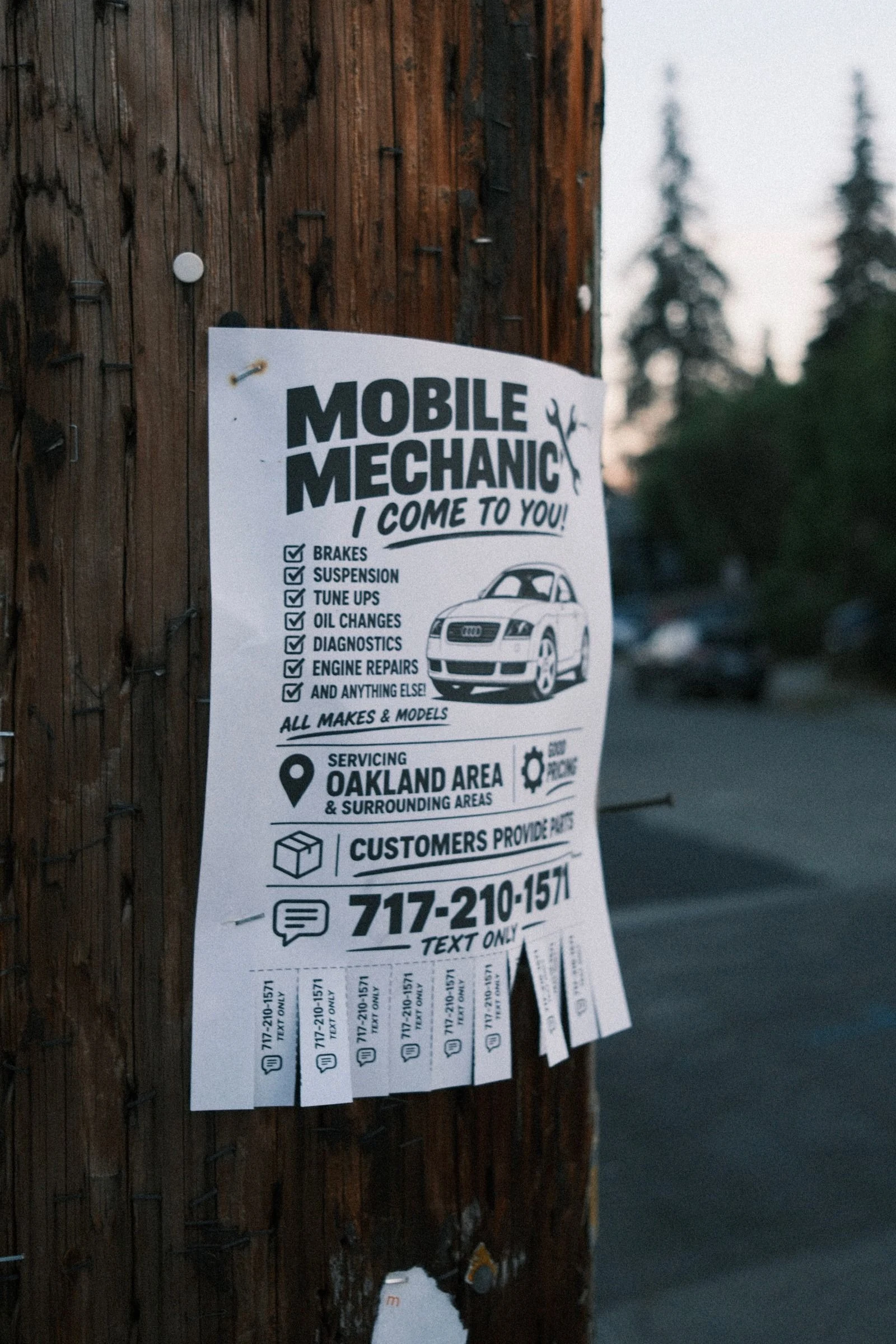 Paper flyer stapled to a weathered wooden utility pole advertising MOBILE MECHANIC, I COME TO YOU, servicing the Oakland area, with tear-off tabs along the bottom and a blurred street and tall pines at dusk behind