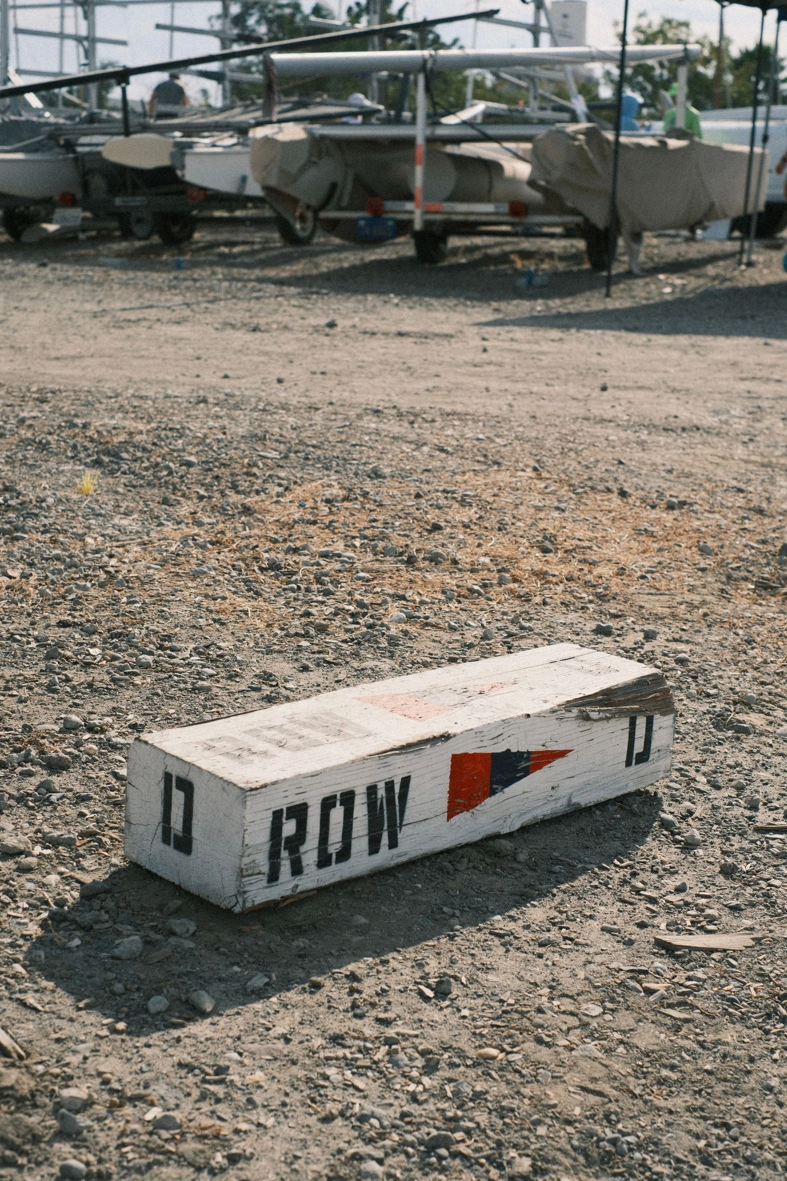 Weathered white wooden block stenciled ROW D with a red and navy pennant, lying on gravel in front of covered sailboats on trailers in a sunny boat yard