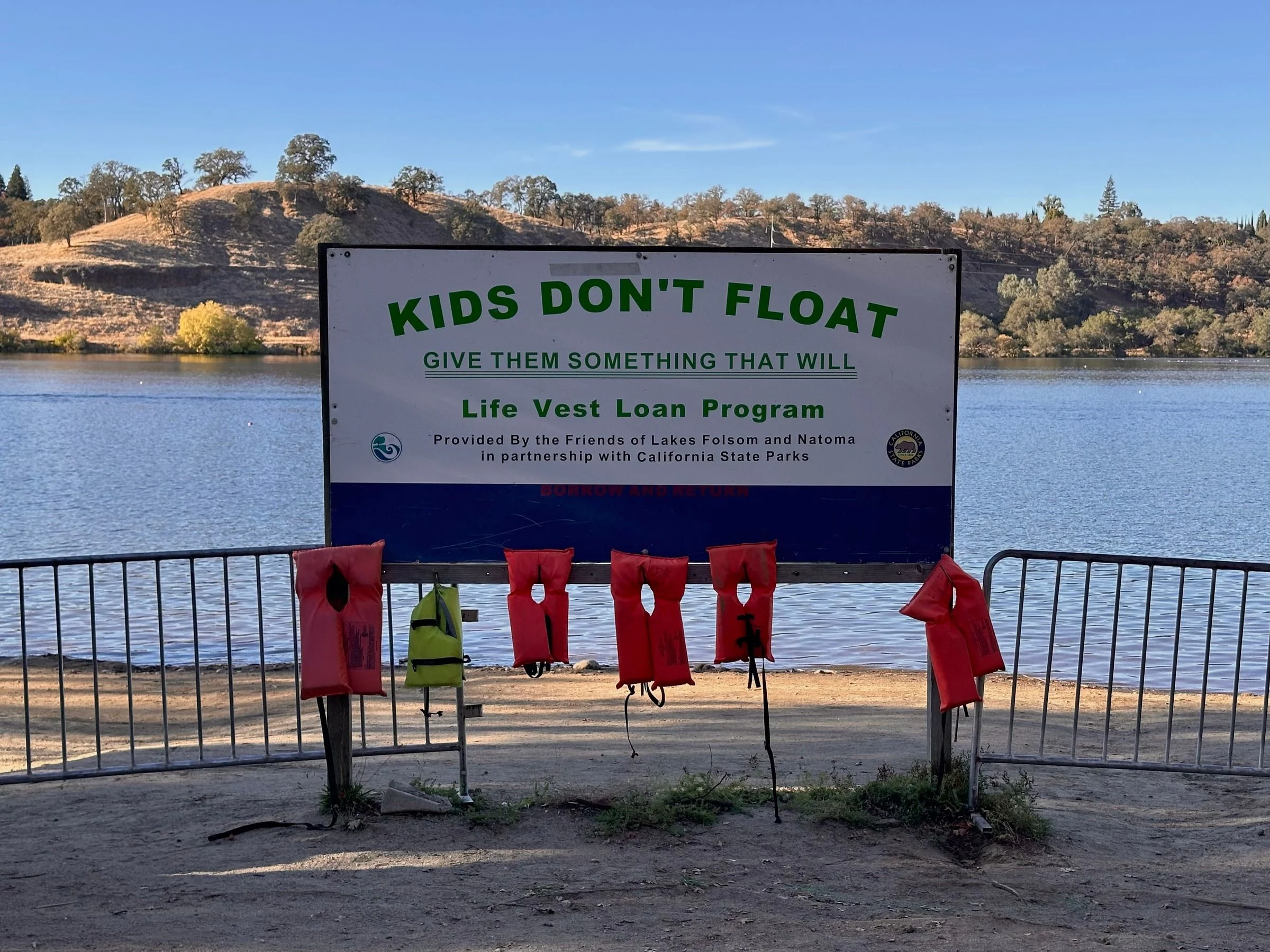 White and blue lakeside sign reading Kids Don't Float Give Them Something That Will Life Vest Loan Program, with six child life vests hanging below it, lake and dry hills behind