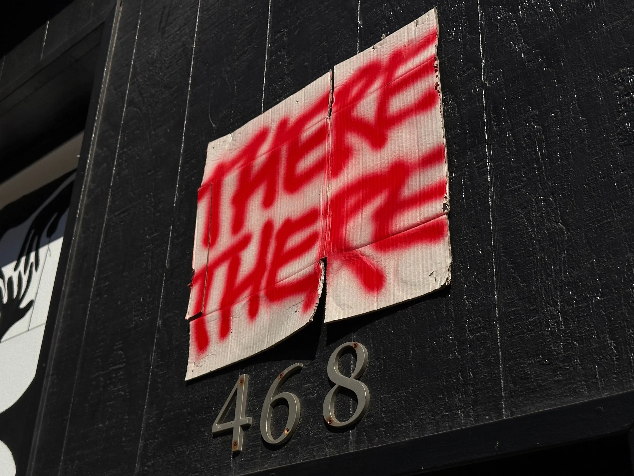 Red spray-painted words THERE THERE on a torn piece of cardboard tacked to a black wooden wall above brushed metal house numbers 468