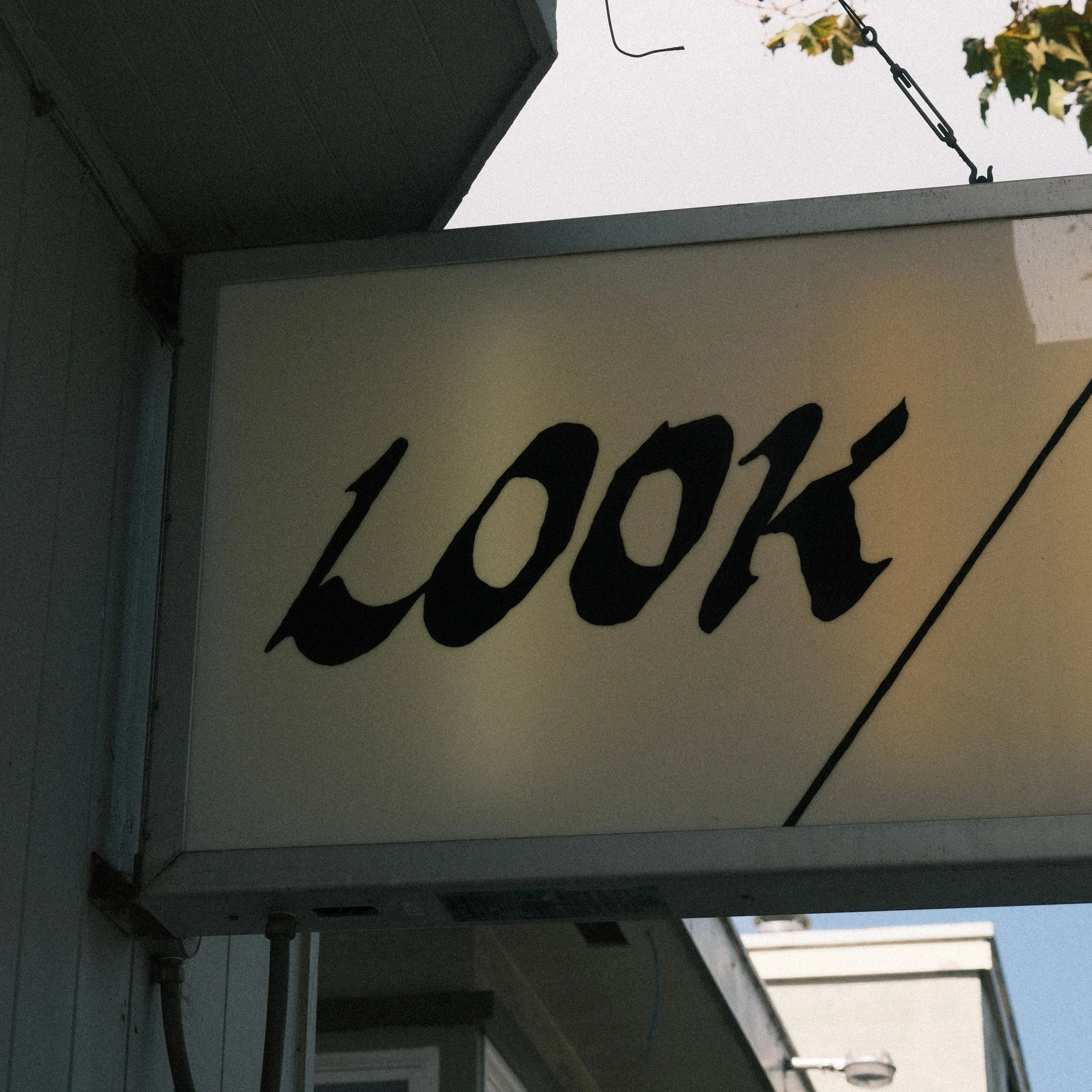 Hand-painted black script spelling LOOK on a cream lightbox sign hanging from a building awning against a pale sky